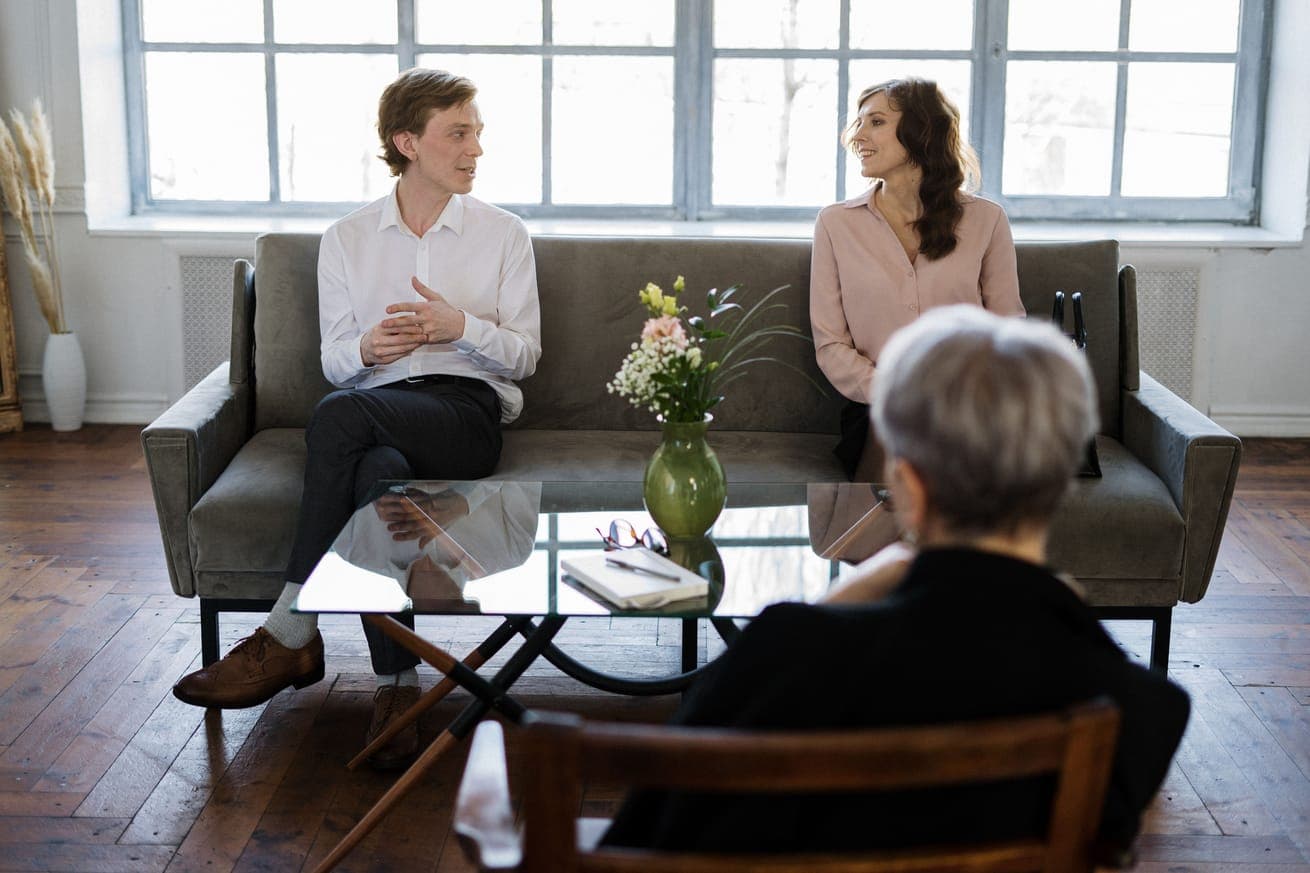 A couple on a grey sofa talking with a grey-haired therapist in a bright room
