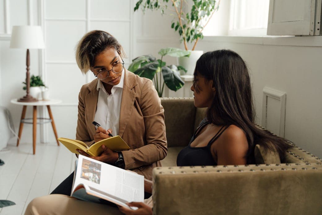 A coach with an open notebook talking with a woman on a sofa