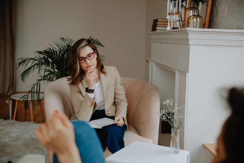 A therapist with glasses in a beige blazer listening from an armchair