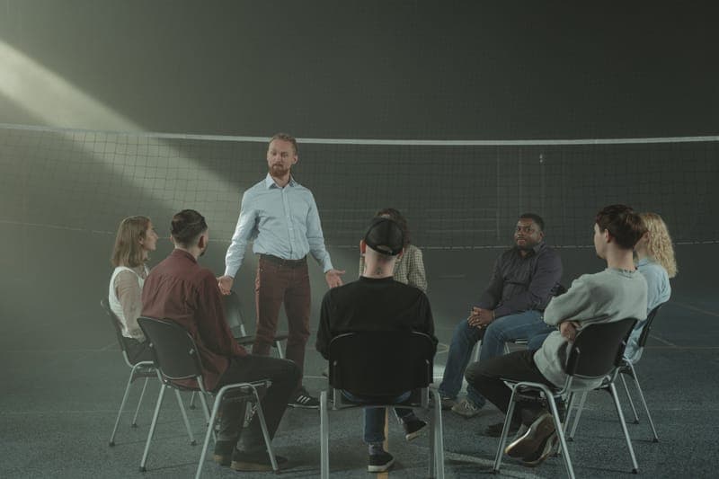 A man standing in the middle of a seated circle in a hall full of light