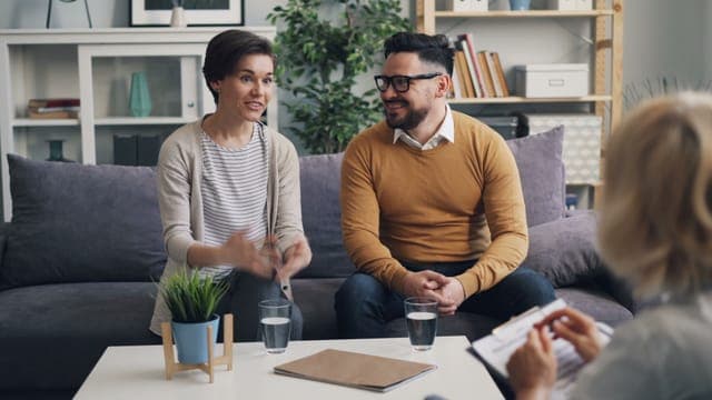 A woman and a bearded man on a sofa talking with a counsellor