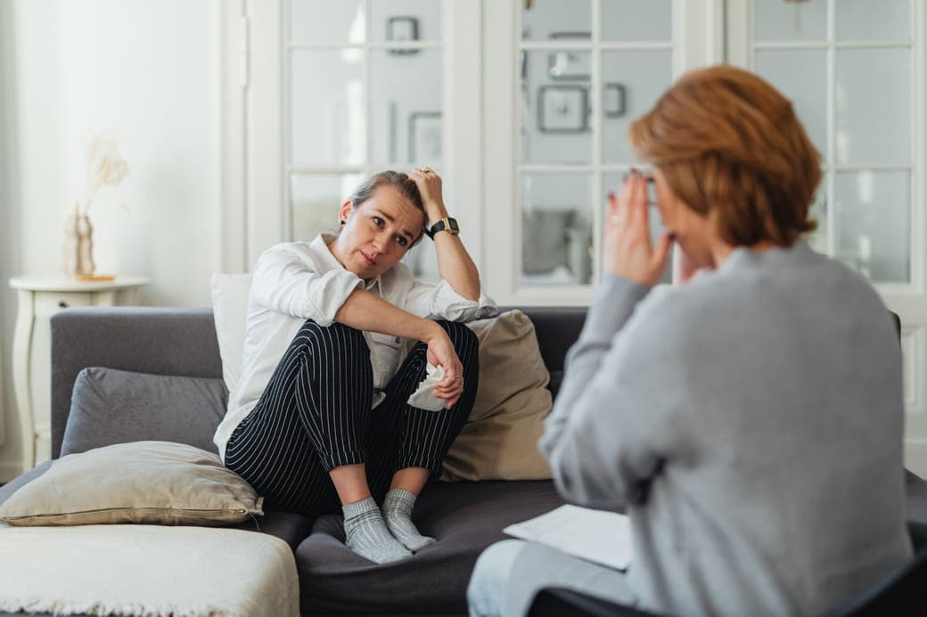 A woman hugging her knee on a sofa, facing a counsellor